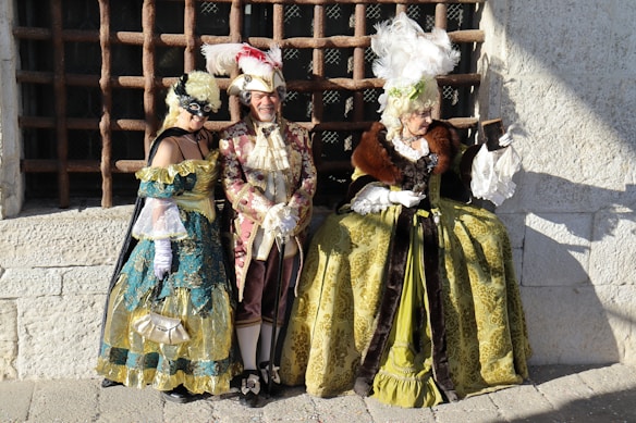 Three individuals are dressed in elaborate historical costumes reminiscent of the Renaissance or Baroque period. They are standing and sitting against a stone building with a large, barred window in the background. The attire includes luxurious fabrics with ornate designs, feathered hats, and a fur cape. One person is wearing a mask, adding a sense of mystery.