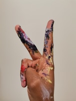 A hand with fingers forming a peace sign is covered in various colors of paint, including black, purple, gold, red, and blue. The paint is splattered and smeared across the fingers, suggesting a creative or artistic activity. The background is a plain off-white color.