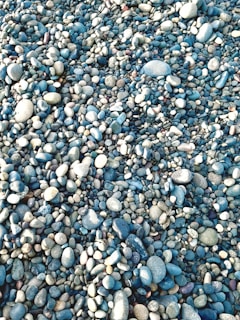 A large assortment of small and medium-sized, smooth stones and pebbles of various shapes and colors are scattered across a surface. The pebbles are predominantly in shades of blue, gray, and white, creating a visually engaging pattern with subtle variations in texture and hue.