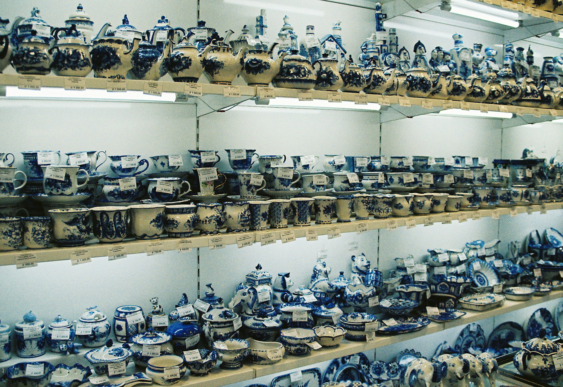 Shelves are filled with various ceramic items, primarily cups, teapots, and plates, all painted in intricate blue and white patterns. The items are arranged in an orderly manner, with price tags visible on each piece. Warm lighting highlights the detailed craftsmanship on the ceramics.
