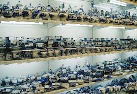 Shelves are filled with various ceramic items, primarily cups, teapots, and plates, all painted in intricate blue and white patterns. The items are arranged in an orderly manner, with price tags visible on each piece. Warm lighting highlights the detailed craftsmanship on the ceramics.