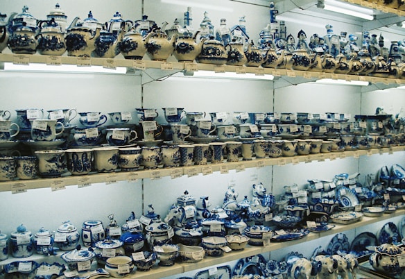 Shelves are filled with various ceramic items, primarily cups, teapots, and plates, all painted in intricate blue and white patterns. The items are arranged in an orderly manner, with price tags visible on each piece. Warm lighting highlights the detailed craftsmanship on the ceramics.