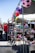 A lively outdoor market scene showcasing a variety of shoes displayed on shelves. Colorful soccer ball decorations hang above the stalls, adding a vibrant touch. People in casual clothing are browsing items in a sunny environment.