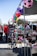 A lively outdoor market scene showcasing a variety of shoes displayed on shelves. Colorful soccer ball decorations hang above the stalls, adding a vibrant touch. People in casual clothing are browsing items in a sunny environment.