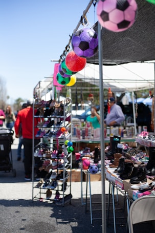 A lively outdoor market scene showcasing a variety of shoes displayed on shelves. Colorful soccer ball decorations hang above the stalls, adding a vibrant touch. People in casual clothing are browsing items in a sunny environment.