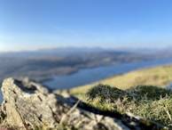 A landscape captures a rocky foreground with grass and moss, overlooking a blurred expanse of water and distant hills under a clear blue sky.