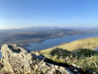 A landscape captures a rocky foreground with grass and moss, overlooking a blurred expanse of water and distant hills under a clear blue sky.