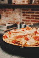 A delicious pizza topped with melted cheese, cherry tomatoes, and slices of prosciutto is placed on a dark tray. The background features a blurred view of a rustic brick wall with kitchen shelves.