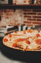 A delicious pizza topped with melted cheese, cherry tomatoes, and slices of prosciutto is placed on a dark tray. The background features a blurred view of a rustic brick wall with kitchen shelves.