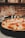A delicious pizza topped with melted cheese, cherry tomatoes, and slices of prosciutto is placed on a dark tray. The background features a blurred view of a rustic brick wall with kitchen shelves.
