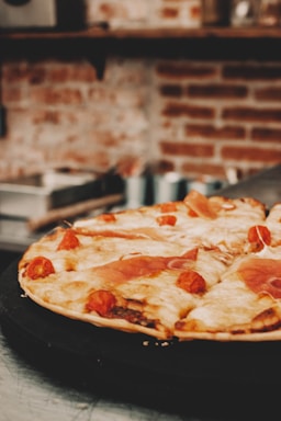 A delicious pizza topped with melted cheese, cherry tomatoes, and slices of prosciutto is placed on a dark tray. The background features a blurred view of a rustic brick wall with kitchen shelves.