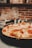 A delicious pizza topped with melted cheese, cherry tomatoes, and slices of prosciutto is placed on a dark tray. The background features a blurred view of a rustic brick wall with kitchen shelves.