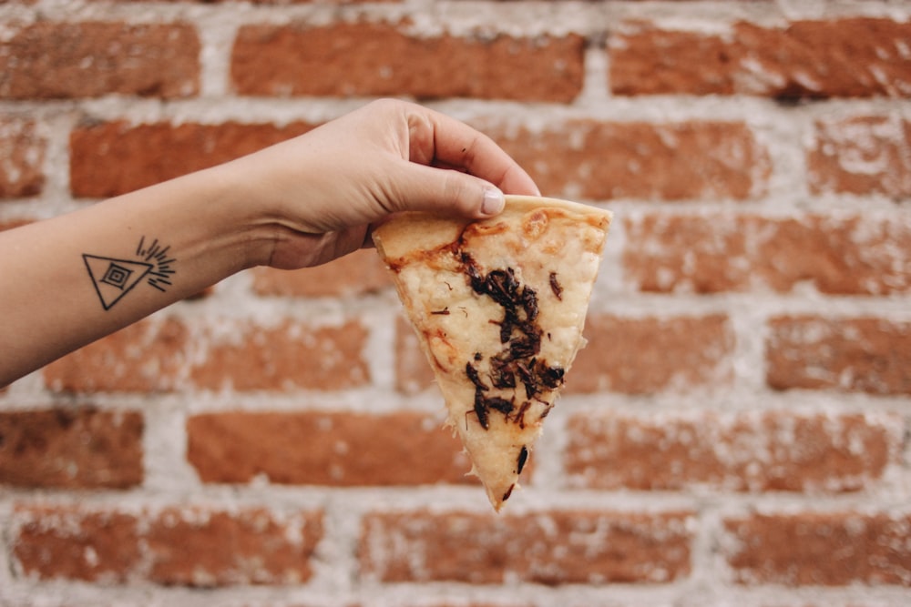 a hand holding a slice of pizza in front of a brick wall