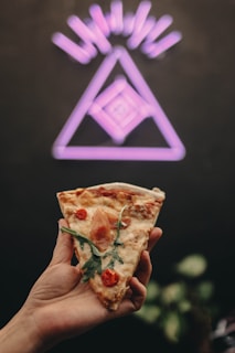 A hand holds a slice of pizza topped with arugula, cherry tomatoes, and a piece of prosciutto. In the background, a neon purple geometric design with a triangle and lines is visible against a dark wall.