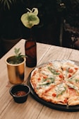 A wooden table displays a freshly baked pizza topped with arugula, tomatoes, and melted cheese, placed on a rustic black plate. Beside it, there is a brass-colored cup filled with a green drink garnished with an herb, and a small black bowl containing a spice mix. A dark glass bottle with a green leaf inserted adds a decorative touch.