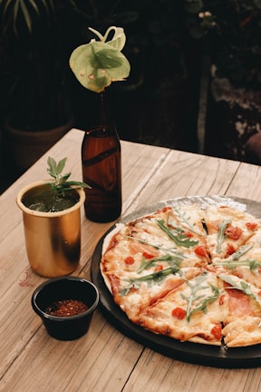 A wooden table displays a freshly baked pizza topped with arugula, tomatoes, and melted cheese, placed on a rustic black plate. Beside it, there is a brass-colored cup filled with a green drink garnished with an herb, and a small black bowl containing a spice mix. A dark glass bottle with a green leaf inserted adds a decorative touch.