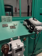 A vintage control panel with teal panels and a rotary dial telephone mounted on it. The setup includes various switches and levers, and there is a sign with instructions. In the background, a fire extinguisher is visible against a white wall.