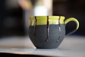 A ceramic mug with a green top and dark matte bottom, adorned with drips of chocolate glaze along the upper edge and handle. It is set on a light-colored surface with a blurred background, highlighting the artistic design of the mug.