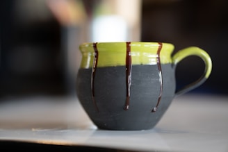 A ceramic mug with a green top and dark matte bottom, adorned with drips of chocolate glaze along the upper edge and handle. It is set on a light-colored surface with a blurred background, highlighting the artistic design of the mug.