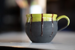 A ceramic mug with a green top and dark matte bottom, adorned with drips of chocolate glaze along the upper edge and handle. It is set on a light-colored surface with a blurred background, highlighting the artistic design of the mug.