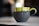 A ceramic mug with a green top and dark matte bottom, adorned with drips of chocolate glaze along the upper edge and handle. It is set on a light-colored surface with a blurred background, highlighting the artistic design of the mug.