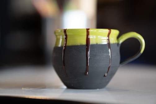 A ceramic mug with a green top and dark matte bottom, adorned with drips of chocolate glaze along the upper edge and handle. It is set on a light-colored surface with a blurred background, highlighting the artistic design of the mug.