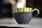A ceramic mug with a green top and dark matte bottom, adorned with drips of chocolate glaze along the upper edge and handle. It is set on a light-colored surface with a blurred background, highlighting the artistic design of the mug.