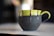 A ceramic mug with a green top and dark matte bottom, adorned with drips of chocolate glaze along the upper edge and handle. It is set on a light-colored surface with a blurred background, highlighting the artistic design of the mug.