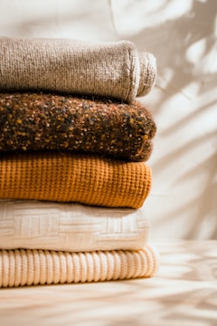 A neatly stacked pile of folded sweaters displayed against a softly lit background. The fabrics vary in texture and color, ranging from beige, brown, mustard yellow to off-white. Soft shadows and warm tones create a cozy atmosphere.