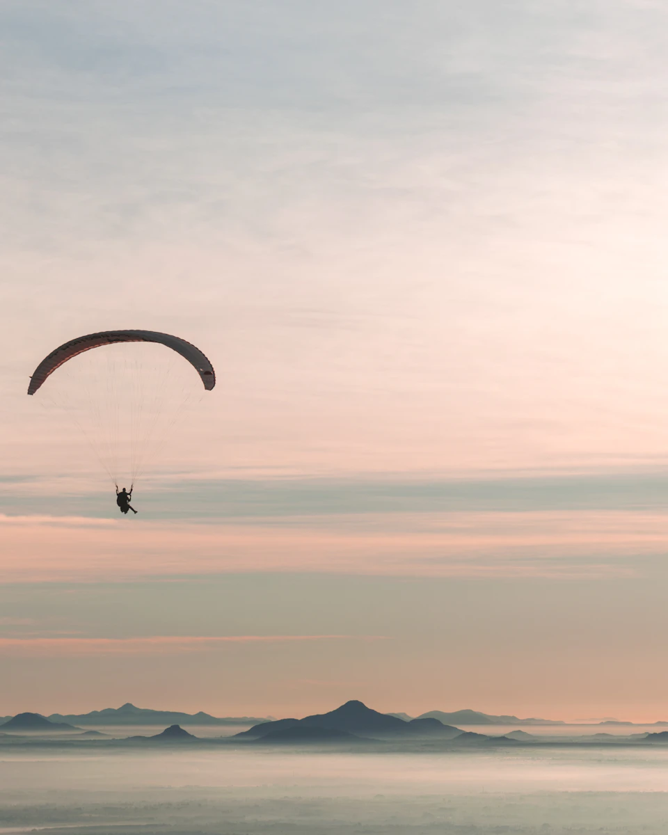 Person in parachute soaring above calm water with billowing parachute against orange sky