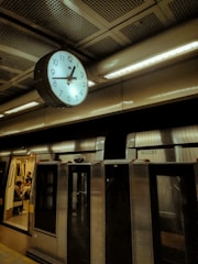 A dimly lit subway station with a large clock showing the time. The clock is mounted on a metallic ceiling. Below the clock, a train is stationed with doors open, revealing passengers seated inside.