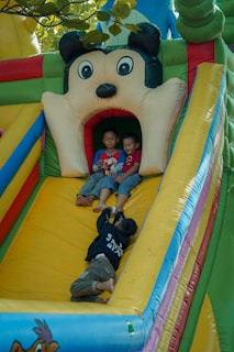 Three children are playing on a colorful inflatable slide. Two children are sitting at the top of the slide, laughing and having fun, while another child is laying on the slide reaching towards them. The slide is designed with a cartoon face at the top, adding to the playful atmosphere.