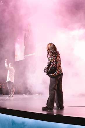 A stage performance with several people partially obscured by pink and white smoke. One person stands in the foreground wearing a patterned jacket and black pants, looking toward the stage. Other performers are visible in the background, and there is a large screen displaying an image of a singer holding a microphone.