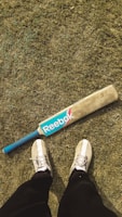 On a grassy field, a cricket bat with a blue Reebok logo and a blue handle is placed on the ground. In the foreground, a pair of feet wearing white sneakers and black pants are visible.