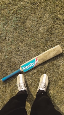 On a grassy field, a cricket bat with a blue Reebok logo and a blue handle is placed on the ground. In the foreground, a pair of feet wearing white sneakers and black pants are visible.