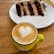 A slice of layered chocolate cake is served on a white plate with a fork. In front of it, there is a yellow mug filled with cappuccino, topped with a heart-shaped latte art. The setting is on a wooden table, creating a cozy and inviting atmosphere.
