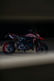 A sleek motorcycle is positioned in a dark setting with minimal lighting, emphasizing its sporty design and red accents. The motorcycle has exposed mechanical parts and is framed by shadows and a blurred foreground.