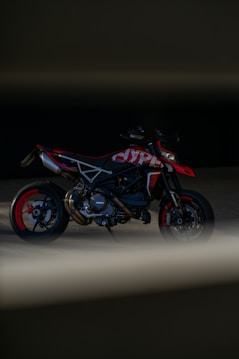 A sleek motorcycle is positioned in a dark setting with minimal lighting, emphasizing its sporty design and red accents. The motorcycle has exposed mechanical parts and is framed by shadows and a blurred foreground.