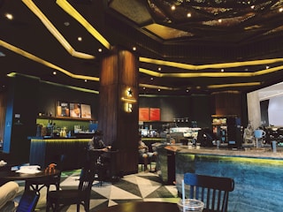 A modern café interior featuring a large counter with coffee machines and a menu board. The lighting is warm and ambient with a patterned ceiling design. Wooden and metal elements dominate the décor. Several tables and chairs are visible, with a person sitting and using a laptop.