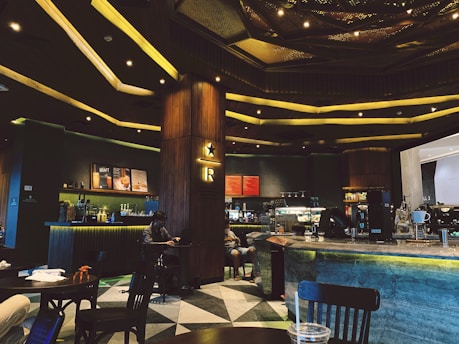 A modern café interior featuring a large counter with coffee machines and a menu board. The lighting is warm and ambient with a patterned ceiling design. Wooden and metal elements dominate the décor. Several tables and chairs are visible, with a person sitting and using a laptop.