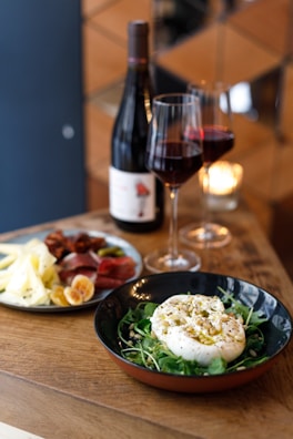 A wooden table holds an elegant spread featuring a plate of assorted cheese, cured meat, and figs next to a bowl of creamy cheese garnished with pine nuts and herbs on a bed of greens. Two glasses of red wine and a bottle are nearby, accompanied by a burning candle, contributing to a cozy and inviting atmosphere.
