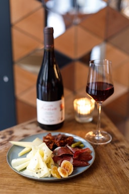 A wooden table is set with a plate of assorted cheeses, cured meats, and dried fruits. Next to the plate, a bottle of red wine and a wine glass filled with wine are positioned. A lit candle in a small glass holder adds a warm ambiance.