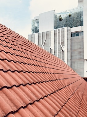 A large, sloped roof covered with red clay tiles, set against a modern, multi-story building with a geometric facade. The building features glass elements and some greenery on an upper balcony, contributing to a contemporary architectural style.