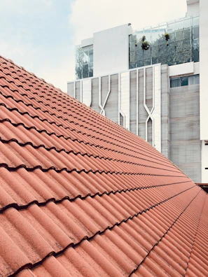 A large, sloped roof covered with red clay tiles, set against a modern, multi-story building with a geometric facade. The building features glass elements and some greenery on an upper balcony, contributing to a contemporary architectural style.