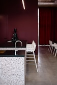 A modern interior space featuring a kitchen counter with a terrazzo surface and a dark countertop. The backdrop consists of a deep maroon wall and matching maroon curtains. A row of minimalist white bar stools line up along the counter. A sleek black coffee machine is placed on the counter. The flooring is a smooth, light grey surface.