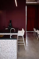 A modern interior space featuring a kitchen counter with a terrazzo surface and a dark countertop. The backdrop consists of a deep maroon wall and matching maroon curtains. A row of minimalist white bar stools line up along the counter. A sleek black coffee machine is placed on the counter. The flooring is a smooth, light grey surface.