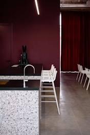 A modern interior space featuring a kitchen counter with a terrazzo surface and a dark countertop. The backdrop consists of a deep maroon wall and matching maroon curtains. A row of minimalist white bar stools line up along the counter. A sleek black coffee machine is placed on the counter. The flooring is a smooth, light grey surface.