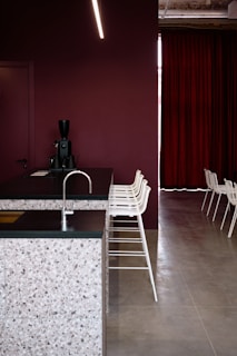 A modern interior space featuring a kitchen counter with a terrazzo surface and a dark countertop. The backdrop consists of a deep maroon wall and matching maroon curtains. A row of minimalist white bar stools line up along the counter. A sleek black coffee machine is placed on the counter. The flooring is a smooth, light grey surface.