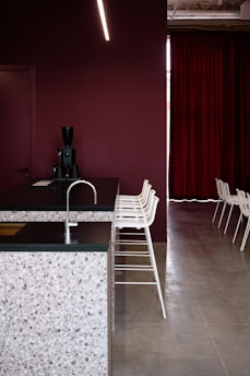 A modern interior space featuring a kitchen counter with a terrazzo surface and a dark countertop. The backdrop consists of a deep maroon wall and matching maroon curtains. A row of minimalist white bar stools line up along the counter. A sleek black coffee machine is placed on the counter. The flooring is a smooth, light grey surface.