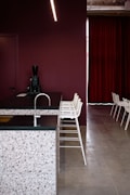 A modern interior space featuring a kitchen counter with a terrazzo surface and a dark countertop. The backdrop consists of a deep maroon wall and matching maroon curtains. A row of minimalist white bar stools line up along the counter. A sleek black coffee machine is placed on the counter. The flooring is a smooth, light grey surface.